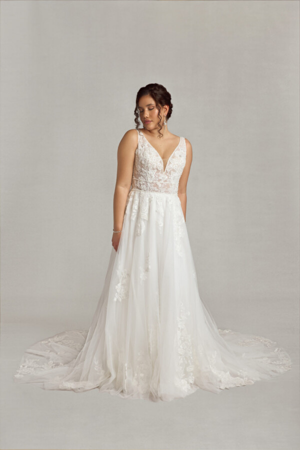 Bride to be wearing a lace wedding dress with a v cut neckline and lace bodice. There are floral decals throughout the different layers of the skirt.