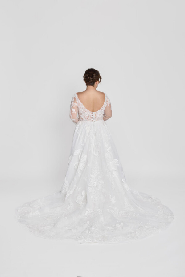 Plus size model wearing a lace sleeve wedding dress with a scoop back and floral detailing on the skirt.