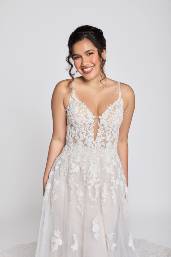 Smiling bride to be wearing a spaghetti strap wedding dress with floral decals on the see through bodice that flow onto the layered, tulle skirt.