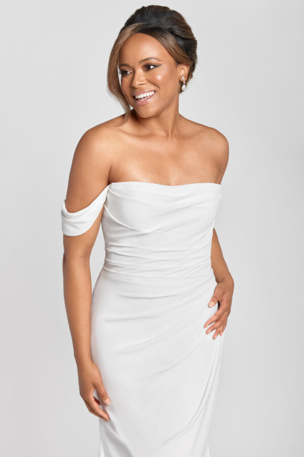 A smiling bride to be wearing an off the shoulder sleeve wedding dress with a ruched bodice and simple skirt.