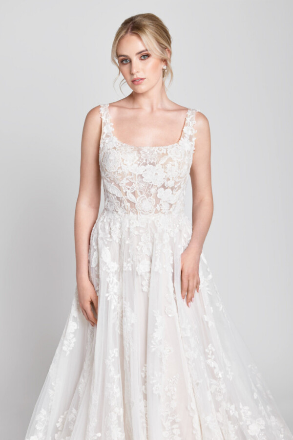 Sweet bride to be wearing a scoop neck wedding dress with floral detailing on the bodice flowing onto the multilayered skirt.