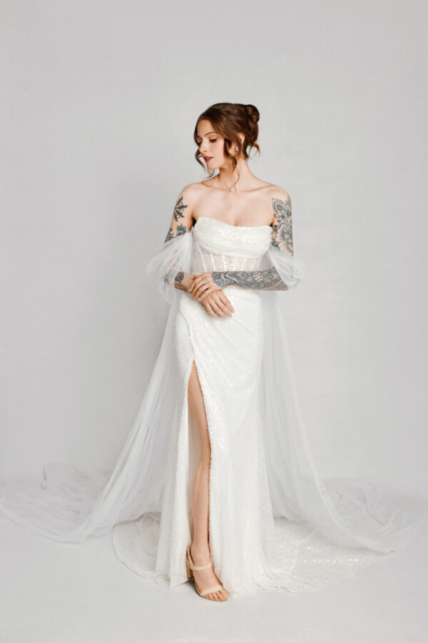 A bride with tattoos in a white fitted lace floral dress with leg slit and arm capes.