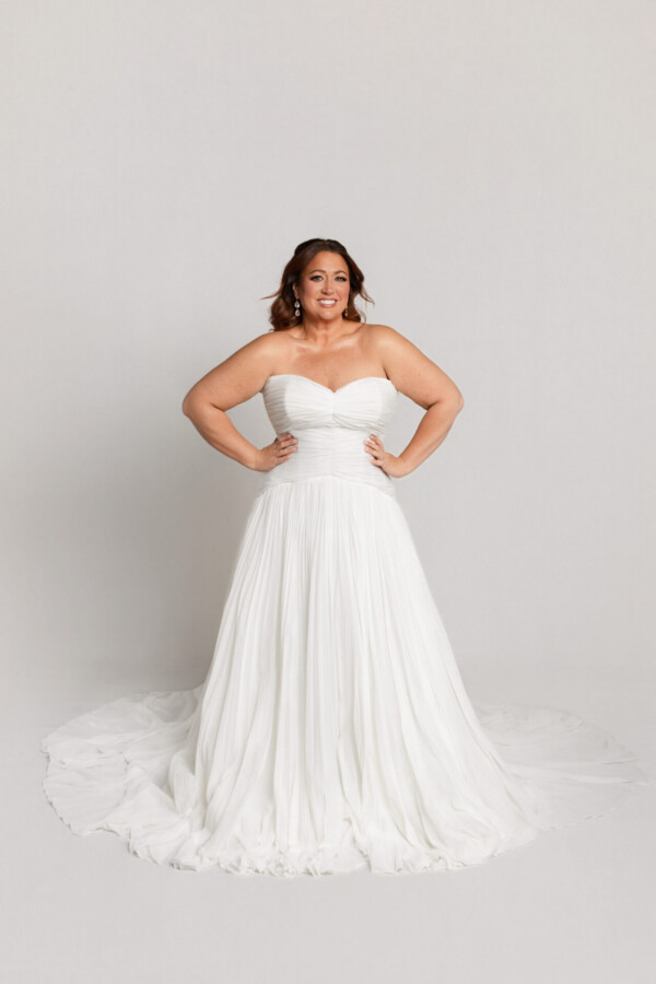 Bride wearing a strapless tulle wedding gown with a sweetheart neckline and full skirt, standing with hands on hips.