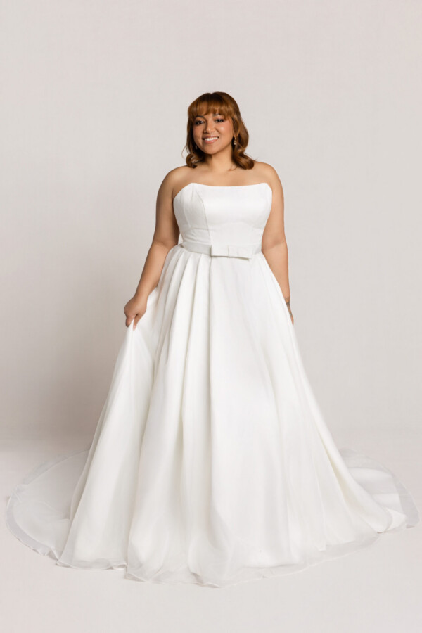 Smiling bride wearing a strapless white wedding gown with a fitted bodice and full skirt, holding the sides of the dress against a neutral background.