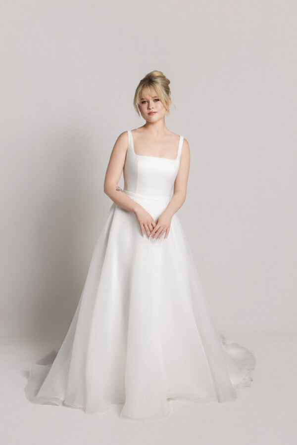Bride wearing a soft tulle wedding dress with thin straps and a full skirt, standing with hands gently clasped.