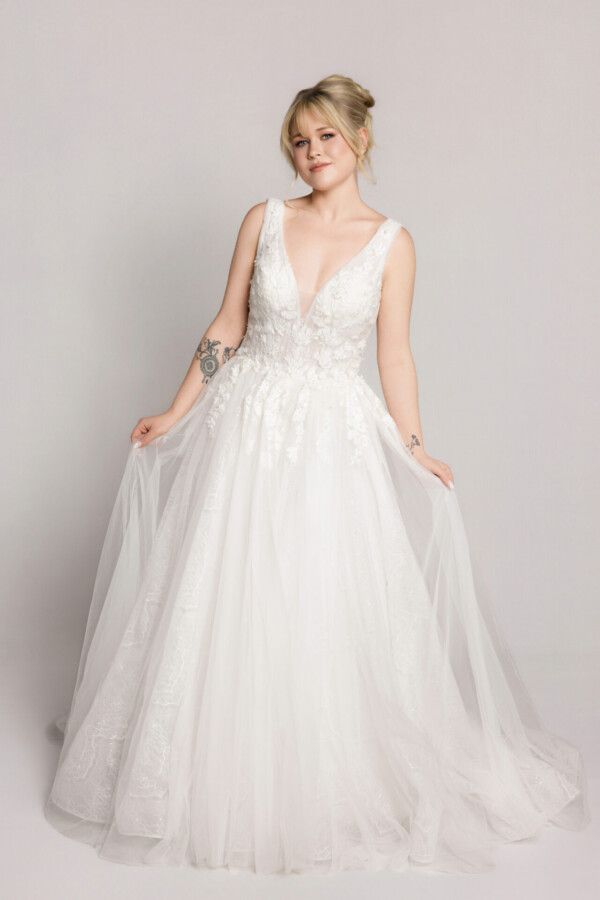 Bride wearing a sleeveless V-neck lace wedding gown with a full tulle skirt, holding the sides of the dress while standing against a neutral background.