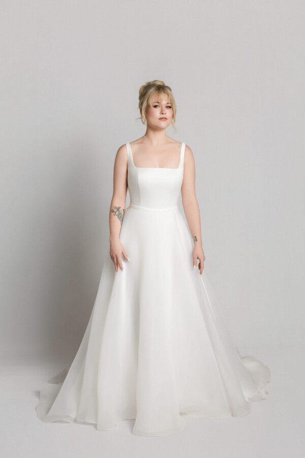 Bride in a soft tulle wedding gown with thin straps and a flowing skirt, standing against a neutral background.
