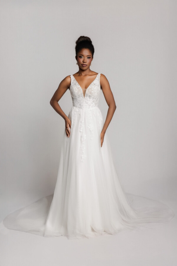 Bride in a sleeveless white wedding gown with a V-neckline and fitted bodice, standing with hands on hips.