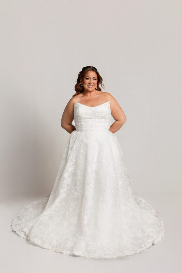 Bride wearing a one-shoulder lace wedding gown with a full skirt and train, standing with hands behind her back against a neutral background.