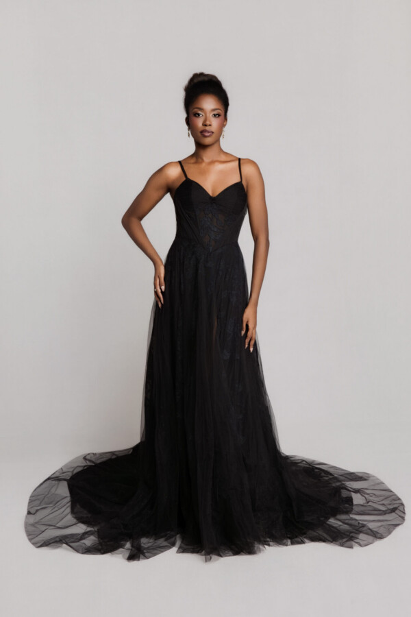 Bride wearing a black wedding gown with thin straps, a fitted bodice, and a sheer tulle skirt with a long train, standing against a neutral background.