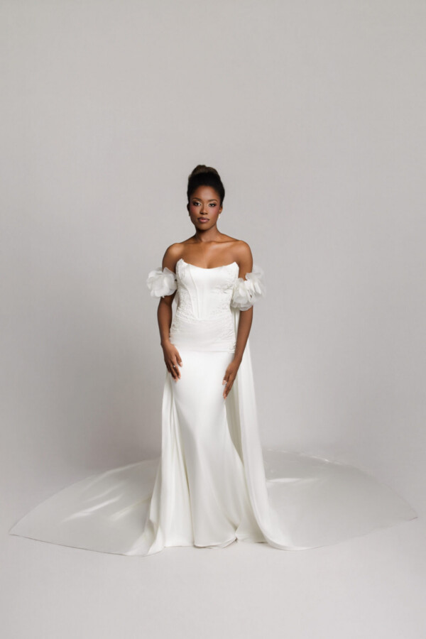Bride in an off-the-shoulder white gown with a fitted silhouette and slight train, standing against a neutral backdrop.