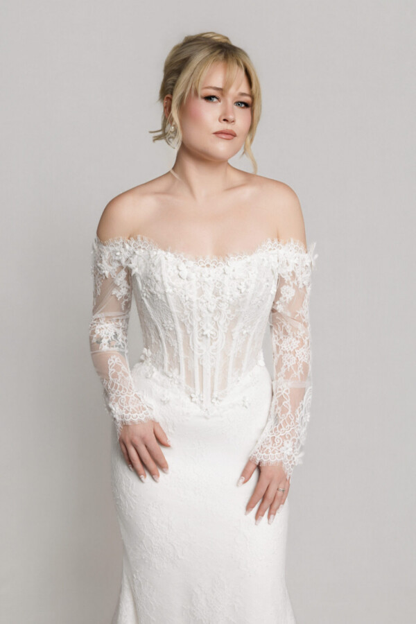 Bride wearing an off-the-shoulder lace wedding gown with long sheer sleeves and a fitted silhouette, posing against a neutral background.