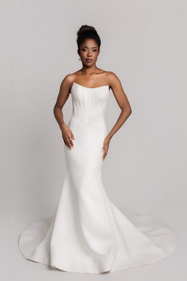 Bride wearing a strapless fitted wedding gown with a slight train, standing against a neutral background.