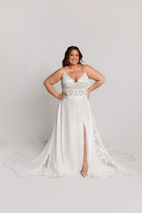 Smiling bride wearing a lace wedding gown with a fitted bodice, thin straps, and a high slit, standing with hands on hips.