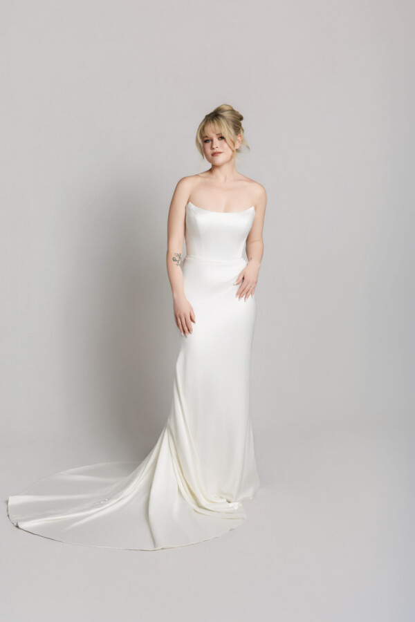 Bride wearing a strapless fitted white wedding gown with a straight neckline and slight train, posing against a neutral background.