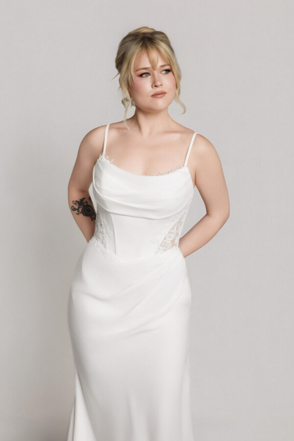 Bride wearing a minimalist white wedding gown with thin straps and a fitted silhouette, posing with hands behind her back.