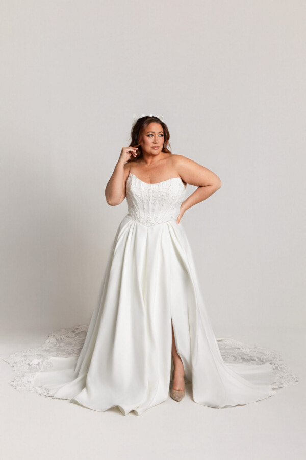 Bride wearing a strapless white gown with a front slit and long train, posing with one hand near her hair against a neutral background.