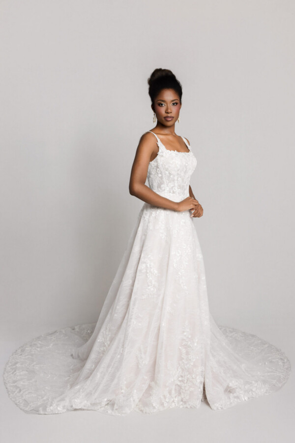 Bride wearing a lace wedding gown with thin straps and a full skirt, standing in a three-quarter pose with a slight train.