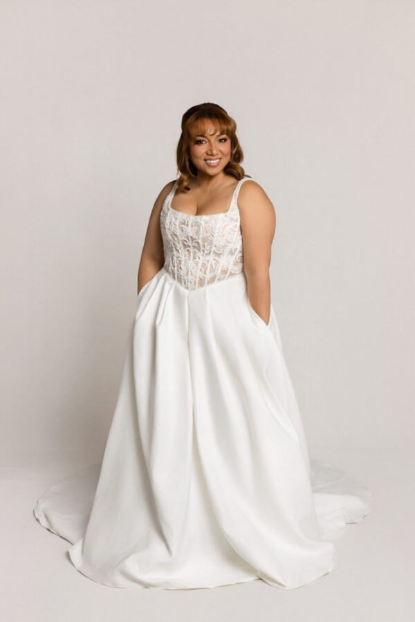 Smiling bride wearing a white wedding gown with a structured bodice and full skirt, standing with hands in pockets.