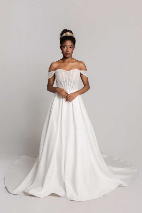 Bride wearing an off-the-shoulder white wedding gown with a full skirt, standing with hands clasped against a neutral background.