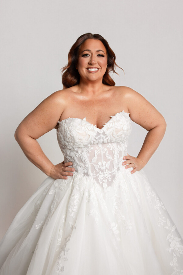Smiling bride wearing a strapless lace wedding gown with a sweetheart neckline and full skirt, standing with hands on hips.