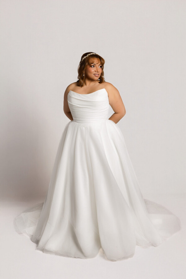 Bride wearing a one-shoulder white wedding gown with a full skirt, standing with hands behind her back against a neutral background.