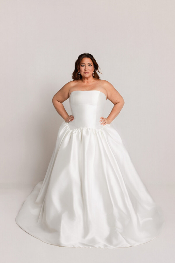 Bride wearing a strapless white ball gown with a fitted bodice and full skirt, standing with hands on hips against a neutral background.