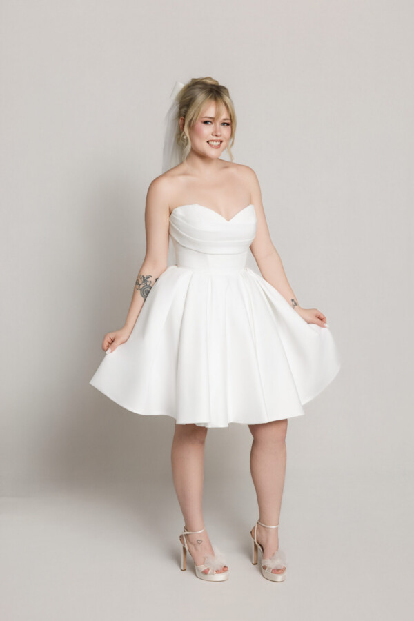 Bride wearing a short strapless white dress with a flared skirt, holding the hem slightly while standing against a neutral background.