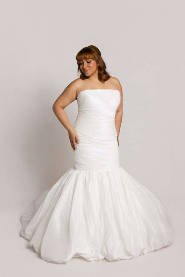 Bride wearing a strapless fitted wedding gown with a mermaid silhouette and flared skirt, posing with hands on hips against a neutral background.