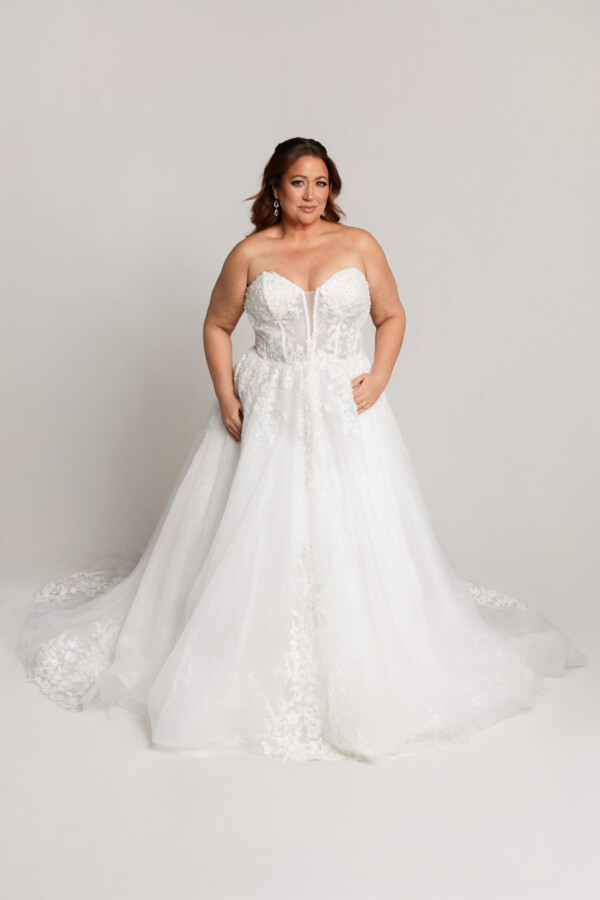 Bride wearing a strapless lace wedding gown with a sweetheart neckline and full tulle skirt, standing against a neutral background.