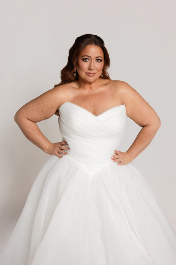 Bride wearing a strapless white wedding gown with a sweetheart neckline and full skirt, standing with hands on hips against a neutral background.