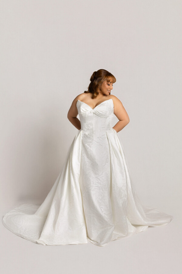 Bride wearing a strapless white wedding gown with a sweetheart neckline and full skirt, posing with hands in pockets and looking to the side.