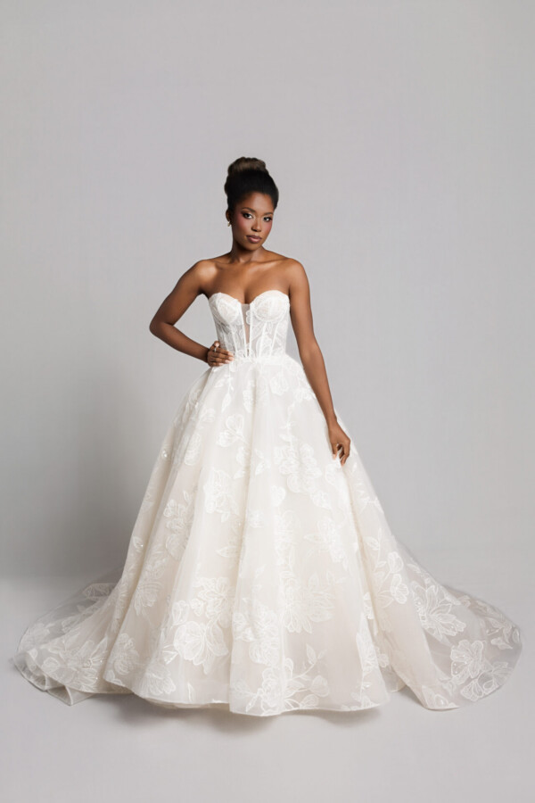 Bride wearing a strapless lace wedding gown with a full skirt and slight train, holding the dress while standing against a neutral background.