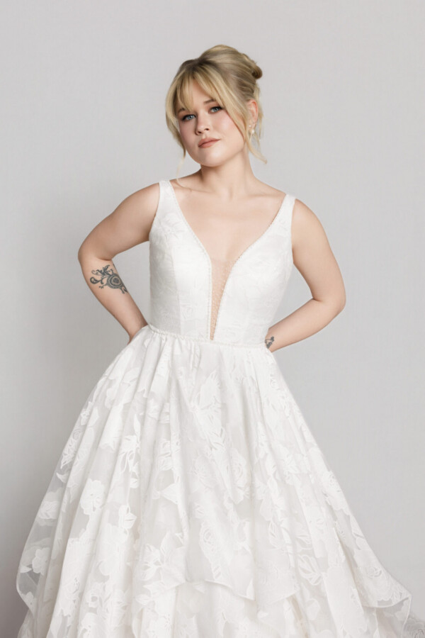 Bride wearing a sleeveless V-neck lace wedding gown with a full skirt, posing with hands on hips.