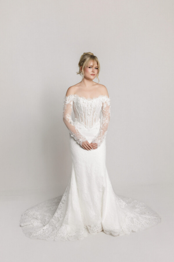 Full-length view of a bride in an off-the-shoulder lace wedding gown with long sleeves and a fitted skirt with a lace train.