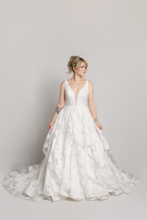 Bride in a sleeveless lace wedding dress with a full skirt, standing against a neutral background.