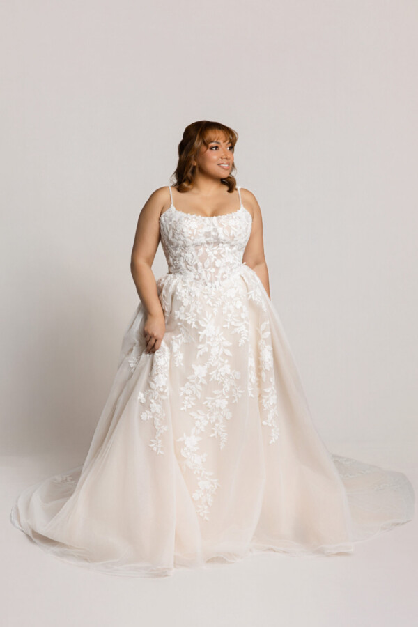 Bride wearing a lace wedding gown with thin straps and a full tulle skirt, holding the sides of the dress while standing against a neutral background.