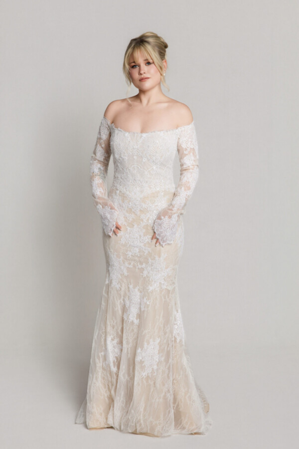 Bride wearing an off-the-shoulder lace wedding gown with long sleeves and a fitted silhouette, standing against a neutral background.