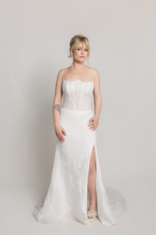Bride in a strapless lace wedding gown with a fitted silhouette and high slit, standing against a neutral background.