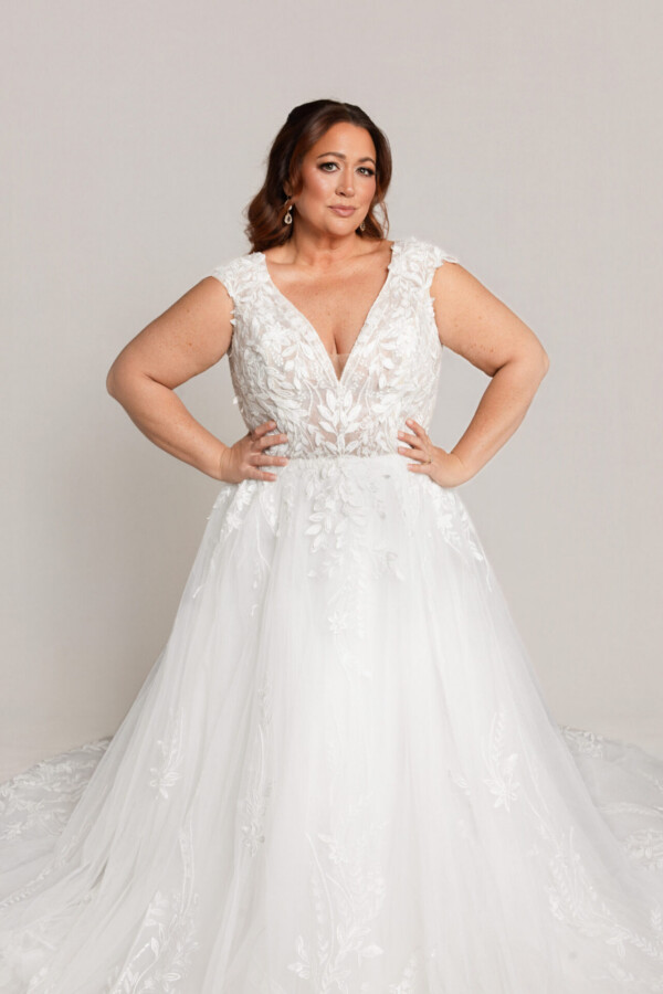 Bride wearing a sleeveless lace wedding gown with a V-neckline and full tulle skirt, standing with hands on hips.