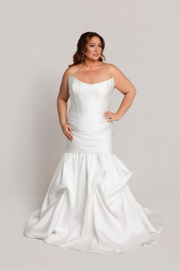 Bride wearing a strapless white wedding gown with a fitted bodice and mermaid-style skirt with ruffled detailing, posing against a neutral background.