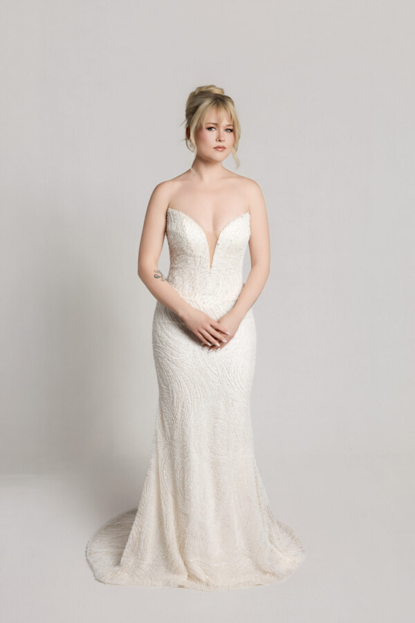 Bride wearing a strapless white wedding gown with a flowing full skirt, holding the sides of the dress while standing against a neutral background.