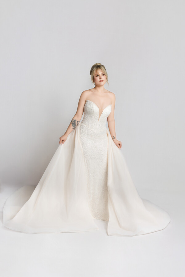 Bride in a strapless fitted wedding gown with a sweetheart neckline and slight train, standing with hands clasped against a neutral backdrop.