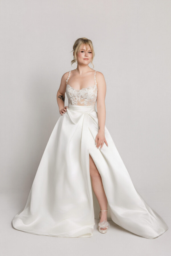 Bride wearing a sleeveless wedding gown with a fitted bodice and full skirt with a high slit, posing against a neutral background.