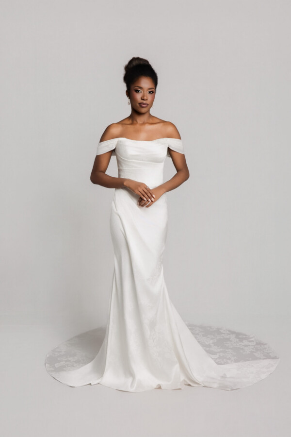 Bride wearing an off-the-shoulder fitted white wedding gown with a mermaid silhouette and slight train, standing against a neutral background.