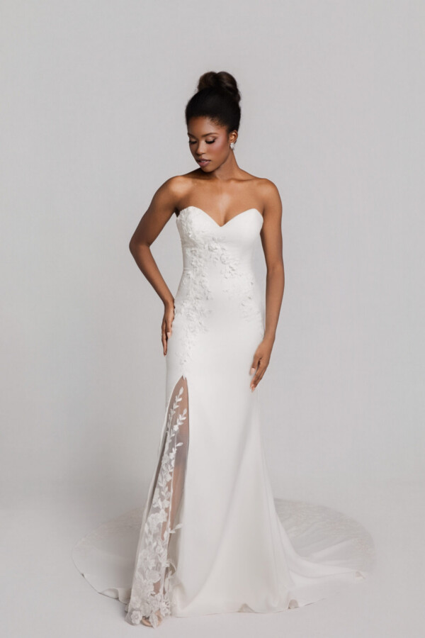 Bride wearing a strapless fitted white wedding gown with a sweetheart neckline and lace side slit, standing against a neutral background.