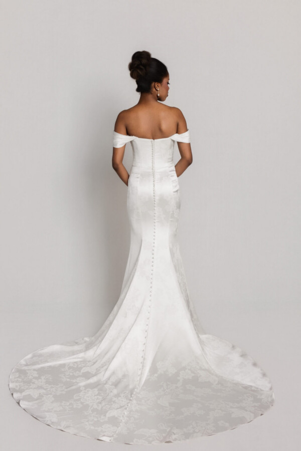 Back view of a bride in an off-the-shoulder mermaid-style wedding gown with a long lace train spread out behind her.