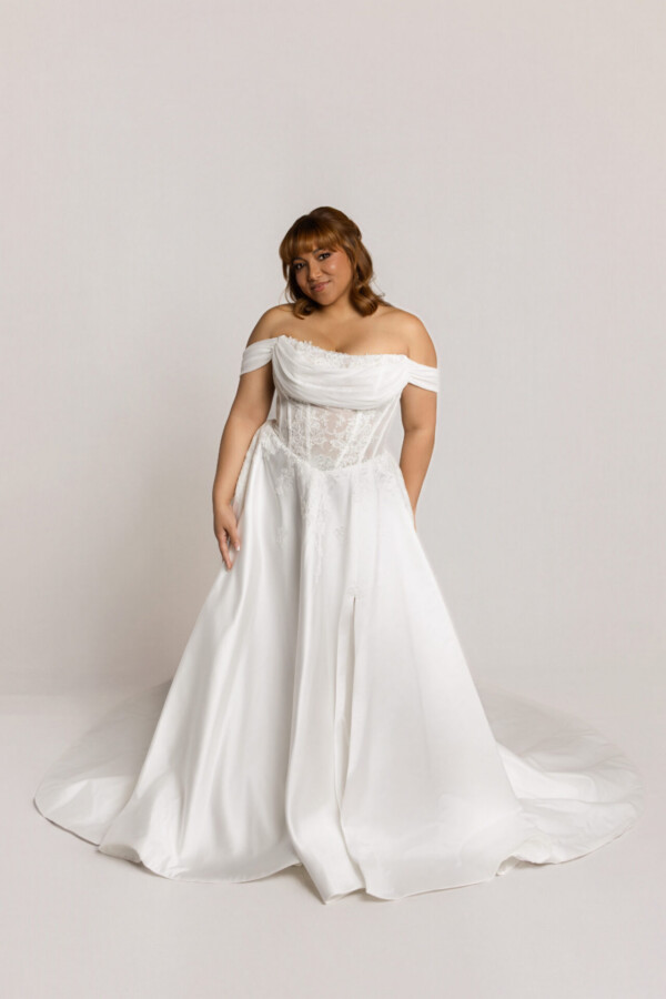 Bride wearing an off-the-shoulder white wedding gown with a full flowing skirt, standing against a neutral background.