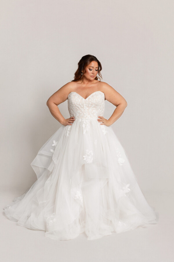 Bride wearing a strapless white wedding gown with a sweetheart neckline and layered tulle skirt, standing with hands on hips against a neutral background.