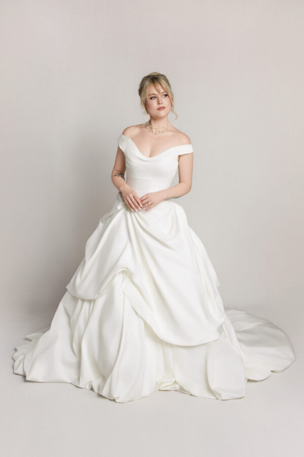 Bride in an off-the-shoulder white wedding gown with a full skirt and soft draping, seated slightly and posing against a neutral background.